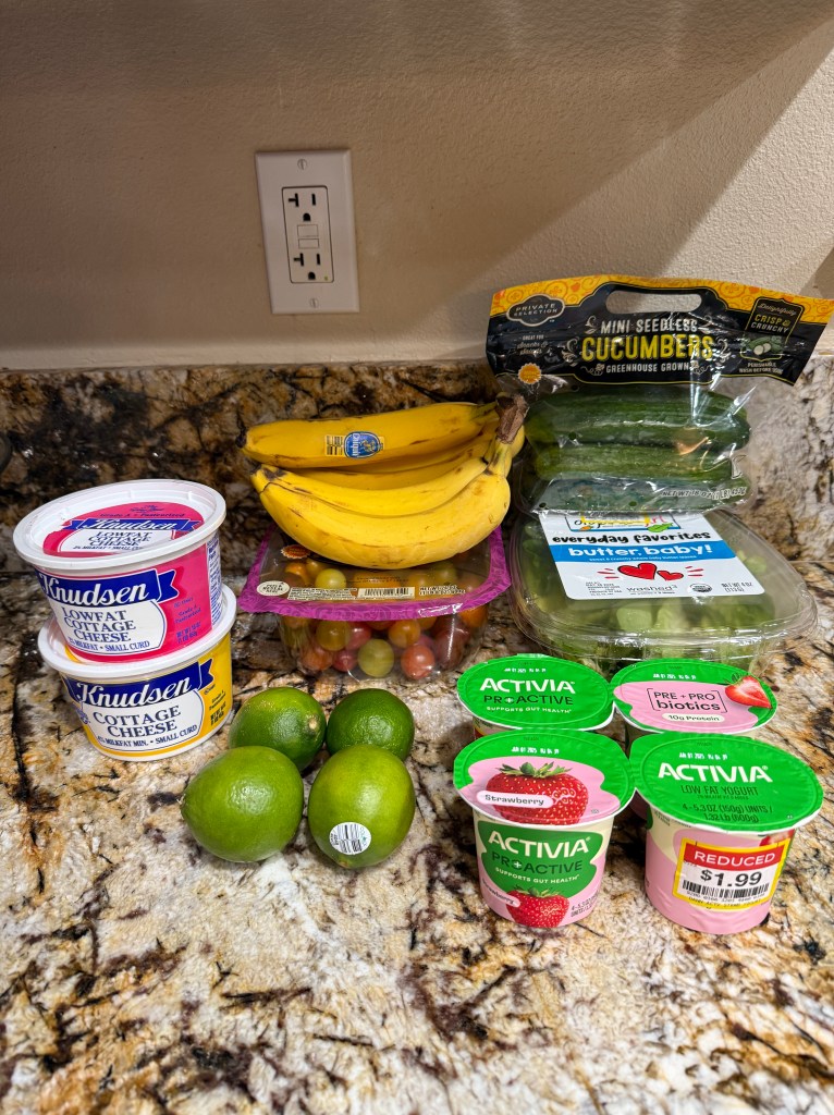 A variety of grocery items on a kitchen counter, including two containers of cottage cheese, a pack of mini seedless cucumbers, a bunch of bananas, three limes, and a pack of cherry tomatoes. Also visible are two cups of Activia yogurt with one marked down to $1.99.