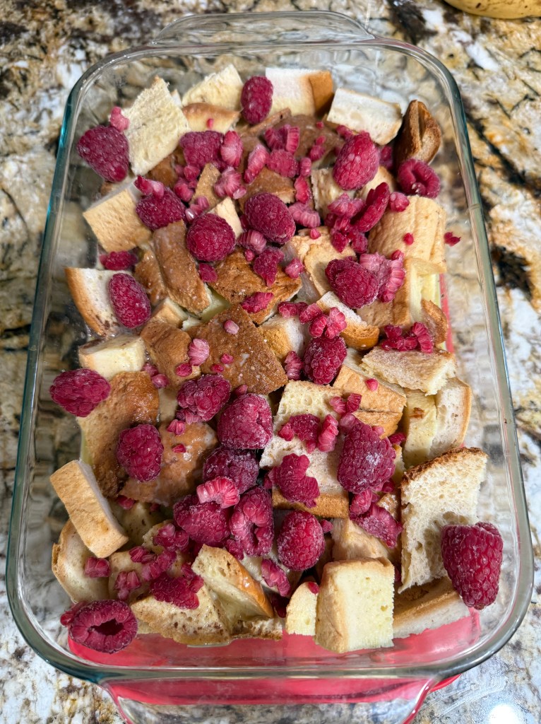 A glass baking dish filled with cubed gluten-free bread and scattered frozen raspberries before baking.