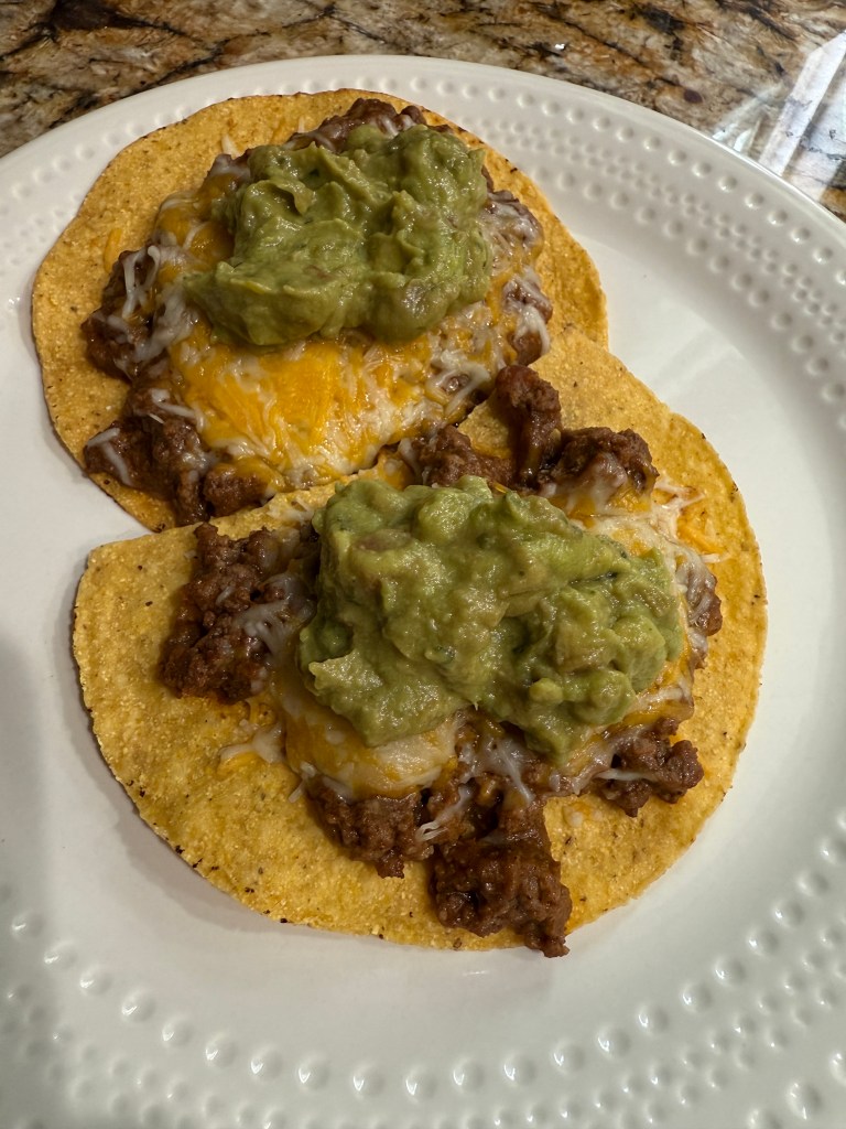 Two tostadas topped with seasoned ground beef, shredded cheese, and guacamole on a white plate.