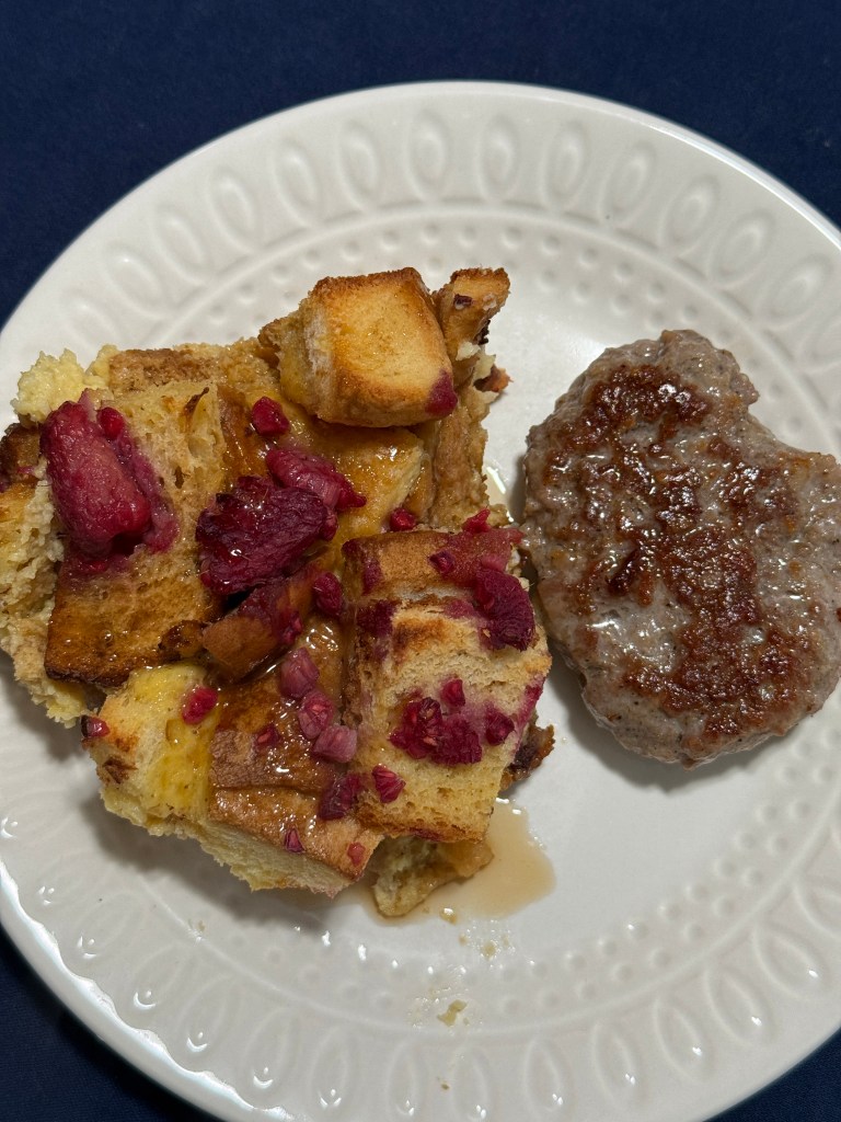 A plate featuring a slice of French toast casserole topped with raspberries and a sausage patty, drizzled with syrup.