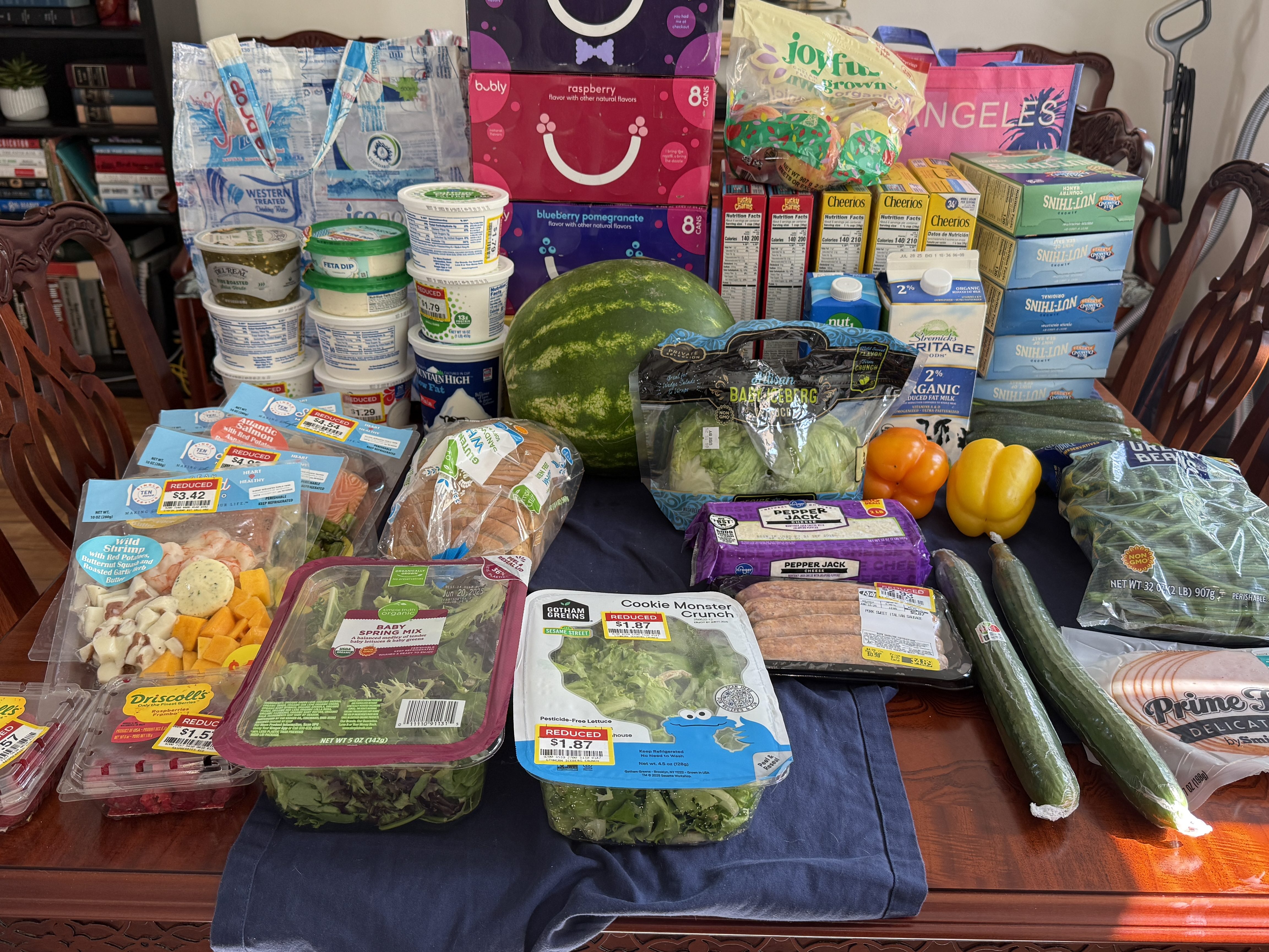 A table full of groceries including various types of cheese, yogurt, produce like lettuce and cucumbers, and bags of beverages. There are sales stickers on some items, showcasing discounts.