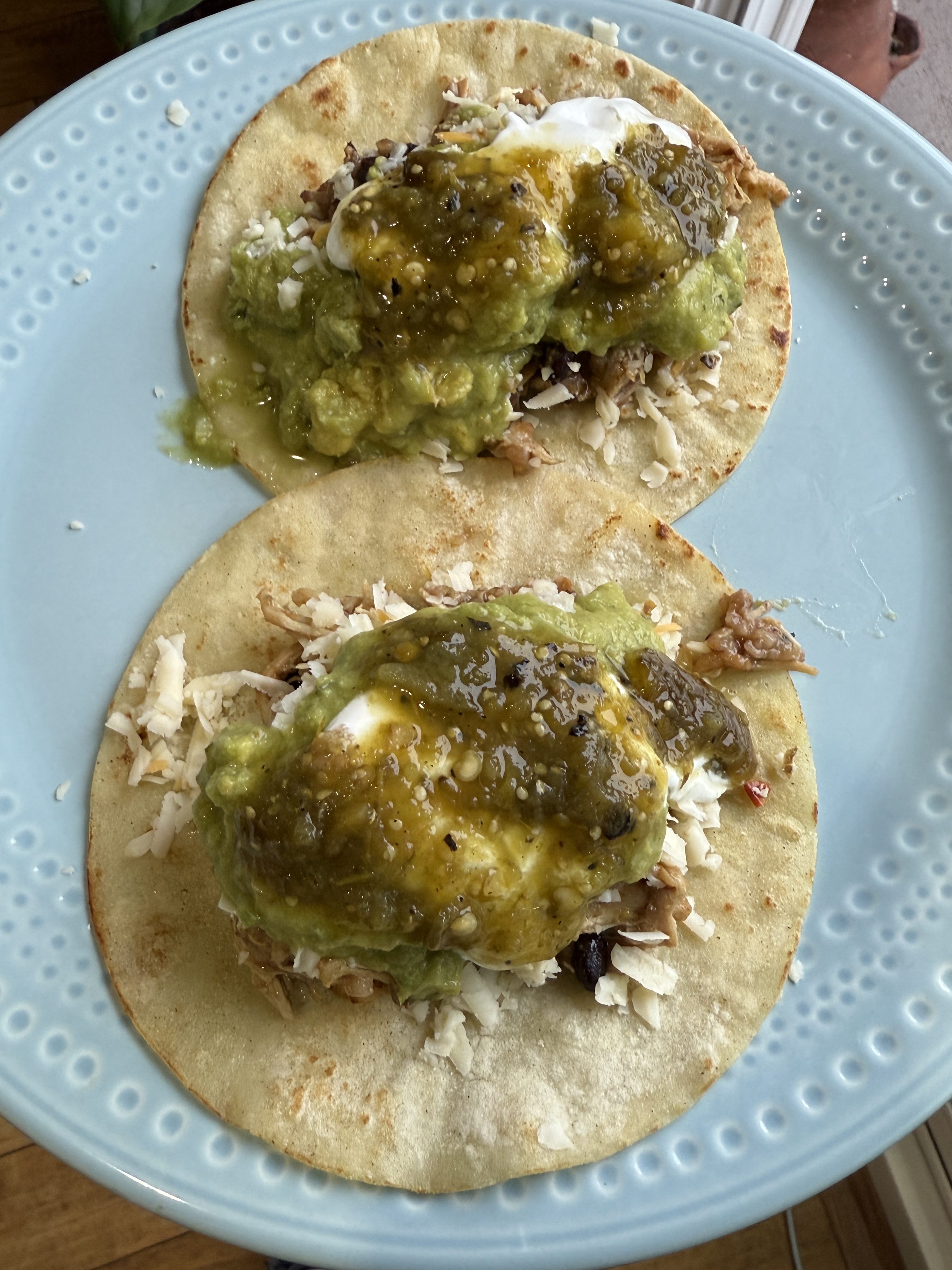Two tacos filled with shredded chicken, black beans, and topped with guacamole, sour cream, and salsa verde, served on a blue plate.