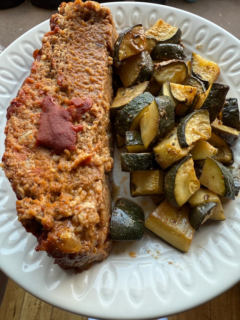 A plate of homemade meatloaf topped with a sauce, accompanied by roasted zucchini pieces.