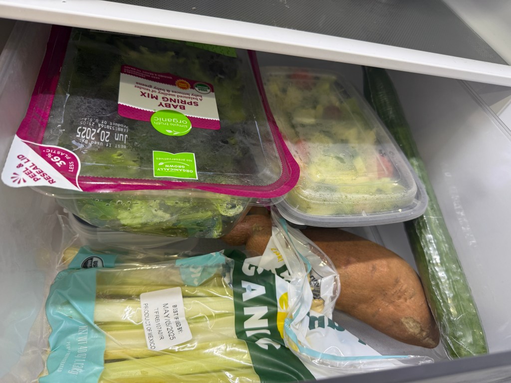 An assortment of fresh vegetables and salad items in a refrigerator, including a bag of salad mix, containers with mixed vegetables, celery, a sweet potato, and a cucumber.