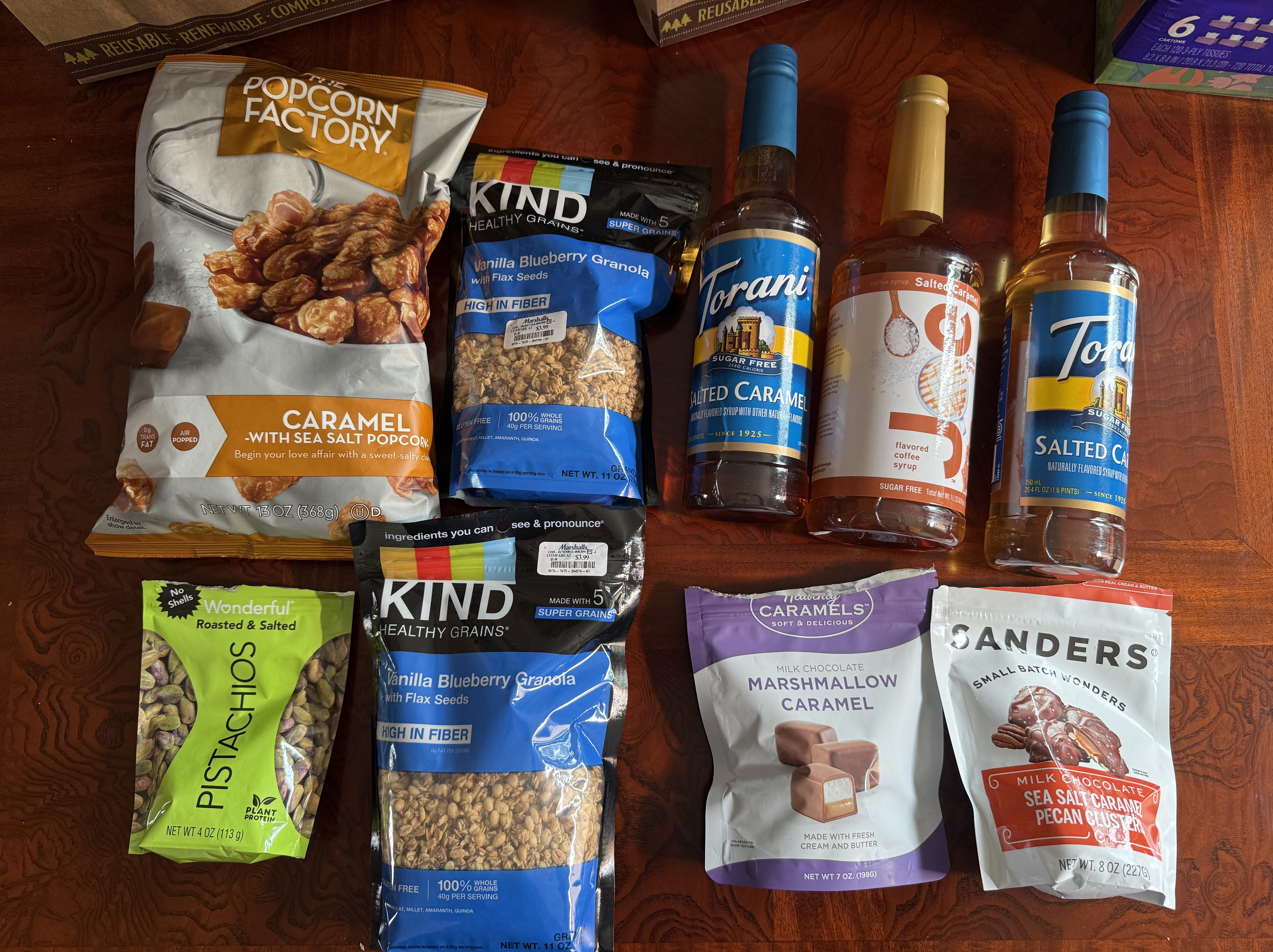 A selection of snacks including caramel popcorn, granola, pistachios, flavored syrup bottles, and chocolate caramels arranged on a wooden surface.