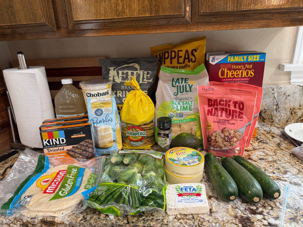 A collection of grocery items on a kitchen countertop, including gluten-free tortillas, snack bars, a cake mix, feta cheese, various vegetables like zucchini and Brussels sprouts, and family-sized cereal boxes.