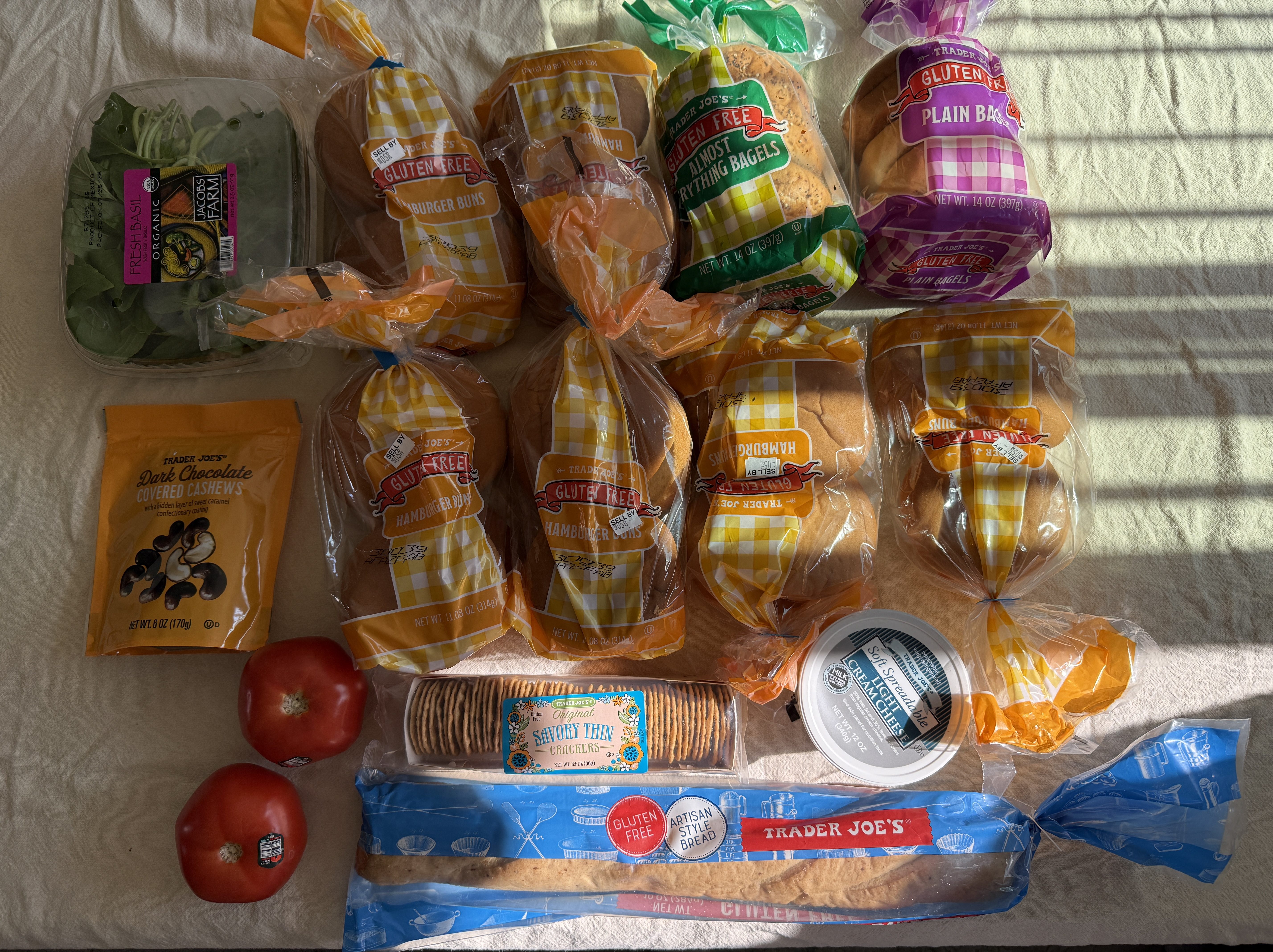 A collection of grocery items on a table, including packages of gluten-free hamburger buns, bagels, a container of fresh spinach, dark chocolate covered cashews, two tomatoes, savory thin crackers, and a container of cream cheese, all arranged neatly with natural lighting.