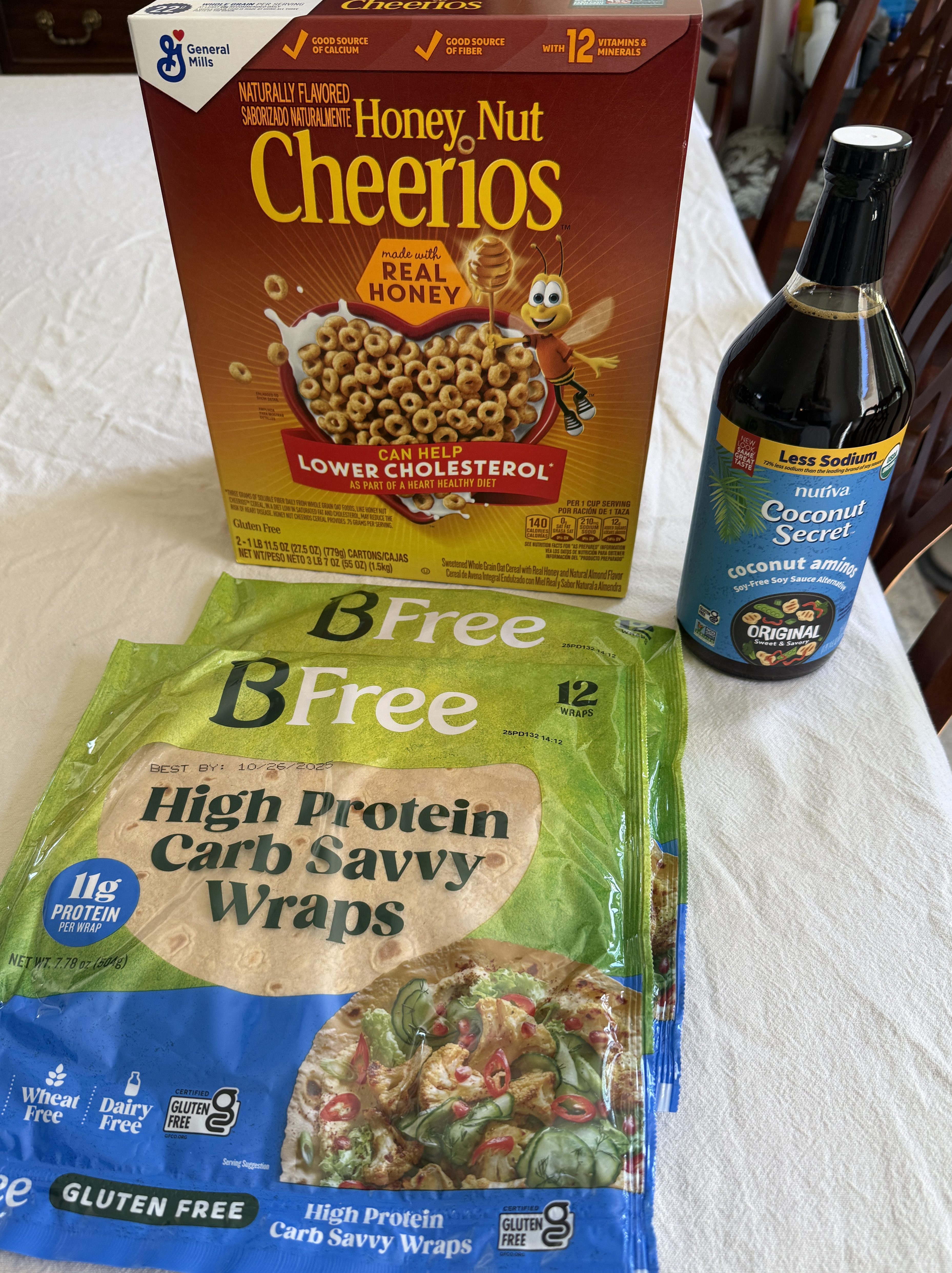 A box of Honey Nut Cheerios resting on a table alongside two packages of BFree High Protein Carb Savvy Wraps and a bottle of Nutiva Coconut Secret Aminos.