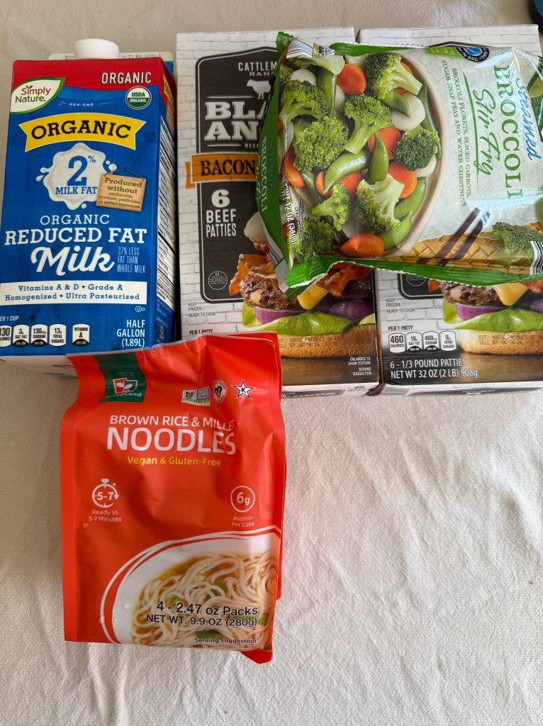A half gallon of organic 2% milk, a pack of bacon beef patties, a bag of steamed broccoli stir fry, and a package of brown rice and millet noodles on a light background.