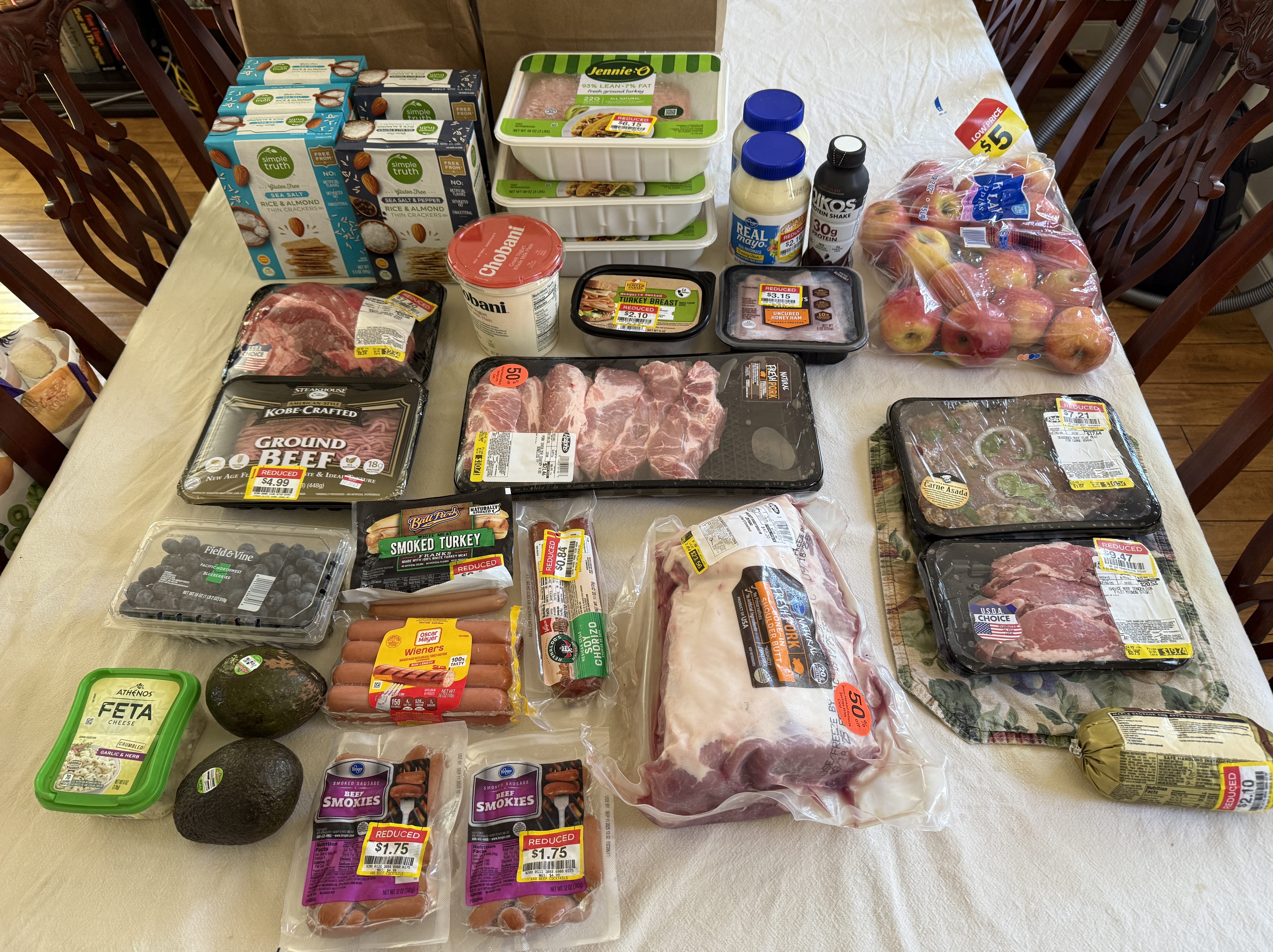 A variety of grocery items displayed on a table, including packs of ground beef, smoked turkey, assorted cheeses, and fresh produce like apples and avocados. Several items are marked with reduced price stickers.